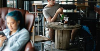 Concentrated male text messaging on cellphone while sitting at table with beverage and photo camera in cafeteria with blurred Asian woman