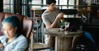 Concentrated male text messaging on cellphone while sitting at table with beverage and photo camera in cafeteria with blurred Asian woman