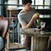 Concentrated male text messaging on cellphone while sitting at table with beverage and photo camera in cafeteria with blurred Asian woman